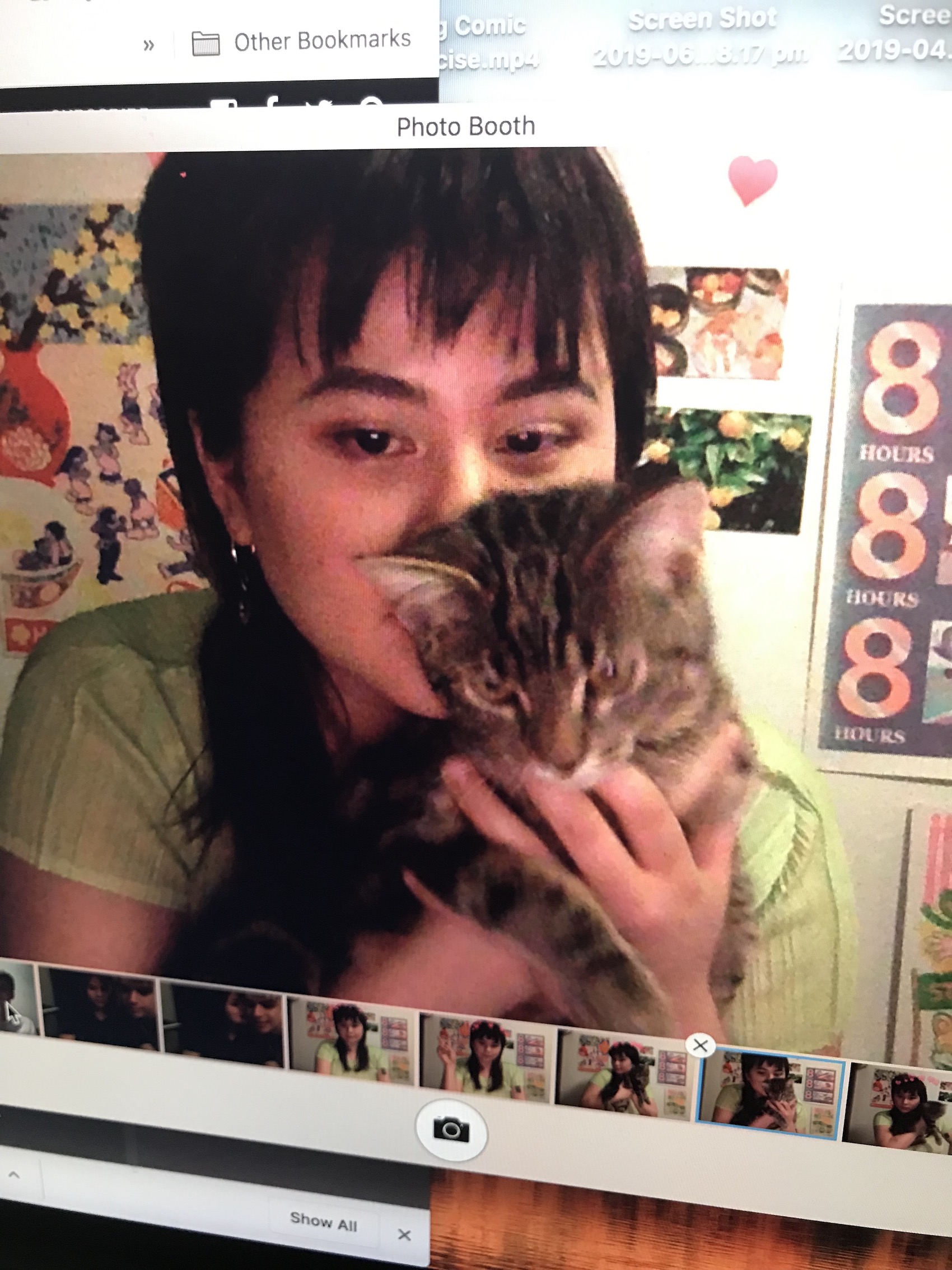 A headshot of an illustrator, Shae San Sim, wearing a short sleeved green top, holding a cat. She has dark hair that sits just past her shoulders. This image is opened and viewed from a Photo Booth window on a computer.